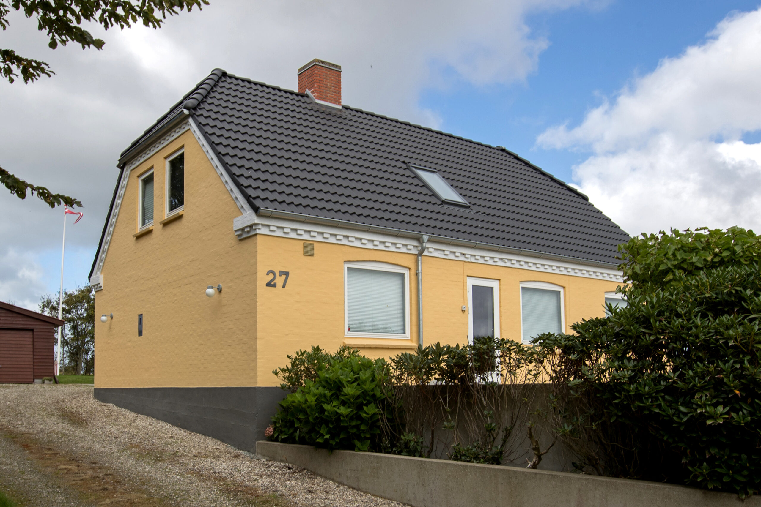 Gult hus med mørkt tegltag og buske